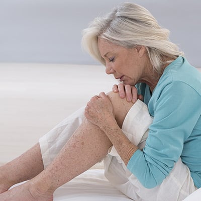 Femme âgée assise sur le sol et se tenant le genou gauche avec une expression de douleur
