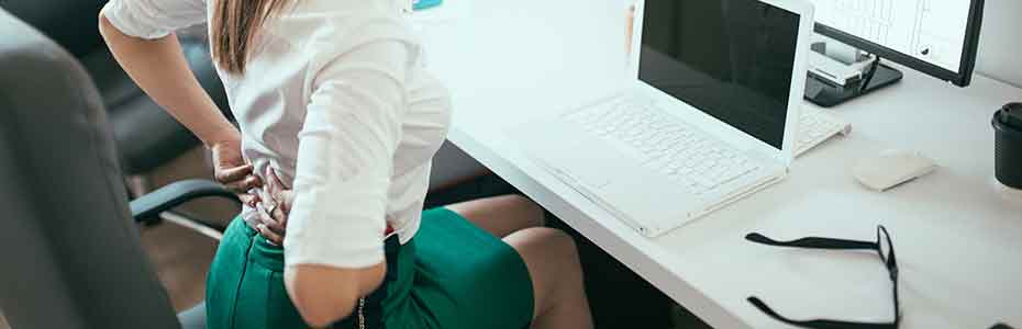 Femme assise à son bureau devant l'ordinateur et qui pose ses deux mains sur le bas de son dos visiblement en douleur